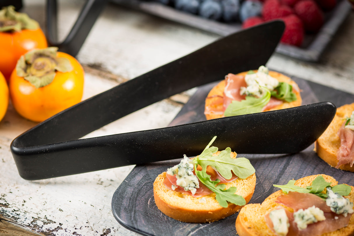 A close-up of crostini topped with blue cheese, arugula, and prosciutto on a dark serving board, with stainless steel tongs resting on top. In the background, persimmons and a tray of assorted berries are visible on a rustic white wooden surface.