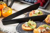 A close-up of crostini topped with blue cheese, arugula, and prosciutto on a dark serving board, with stainless steel tongs resting on top. In the background, persimmons and a tray of assorted berries are visible on a rustic white wooden surface.