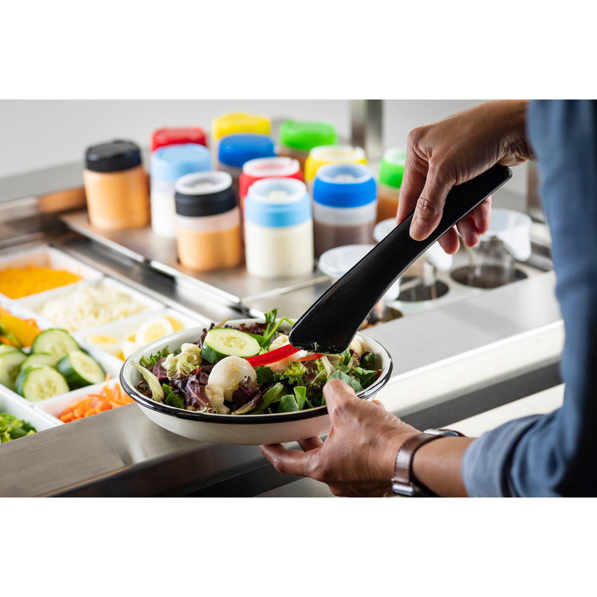 A person uses heat safe tongs to serve sliced red bell pepper onto a bowl of fresh salad. The salad bar in the background features cucumbers, cheese, dressings in plastic bottles, and vibrant vegetables in stainless steel containers.
