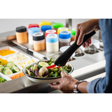 A person uses heat safe tongs to serve sliced red bell pepper onto a bowl of fresh salad. The salad bar in the background features cucumbers, cheese, dressings in plastic bottles, and vibrant vegetables in stainless steel containers.