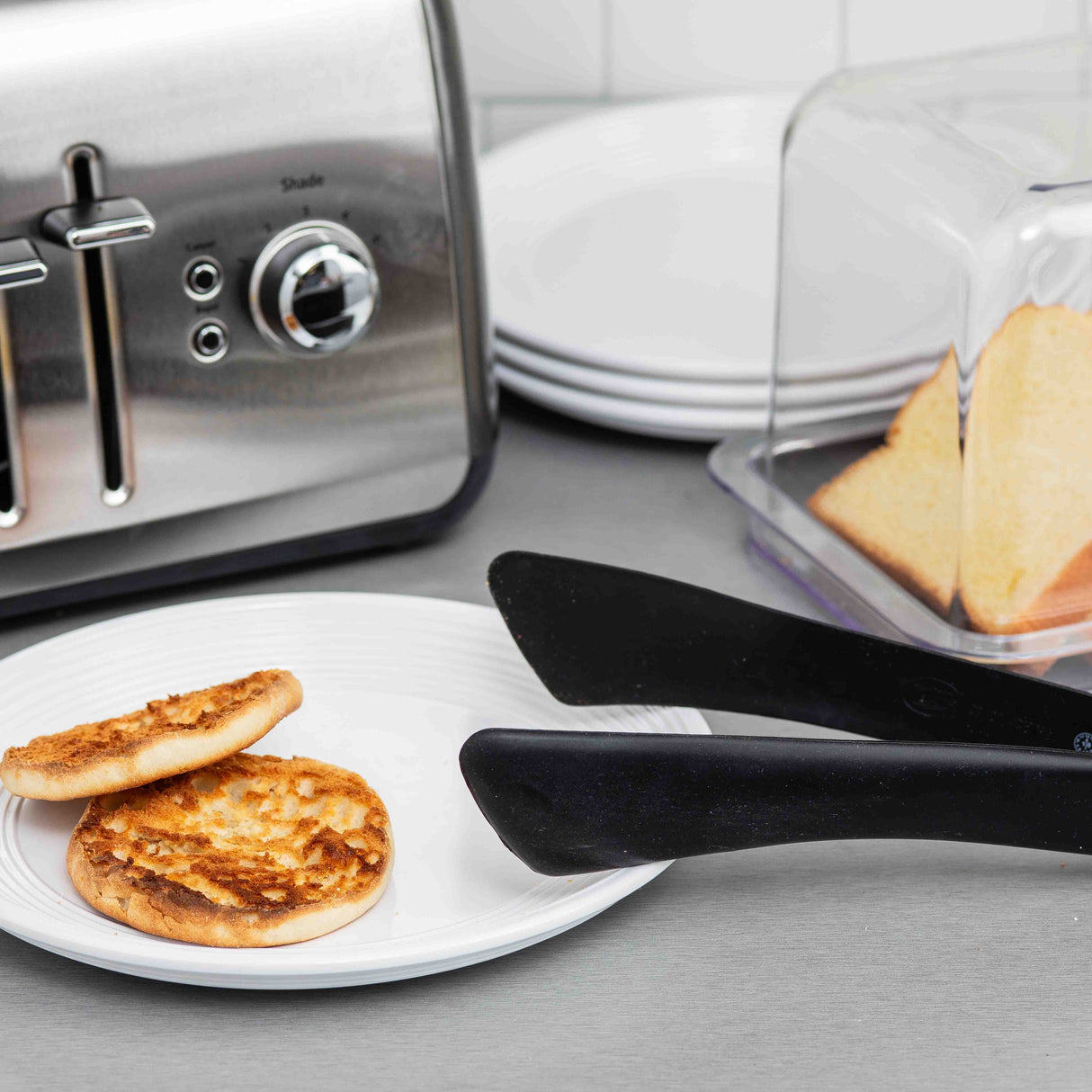 Two toasted English muffin halves sit on a white plate next to heat safe black tongs. Behind them is a stainless steel toaster, a stack of white plates, and a butter dish with two bread slices, all arranged on a gray countertop.