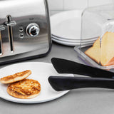 Two toasted English muffin halves sit on a white plate next to heat safe black tongs. Behind them is a stainless steel toaster, a stack of white plates, and a butter dish with two bread slices, all arranged on a gray countertop.