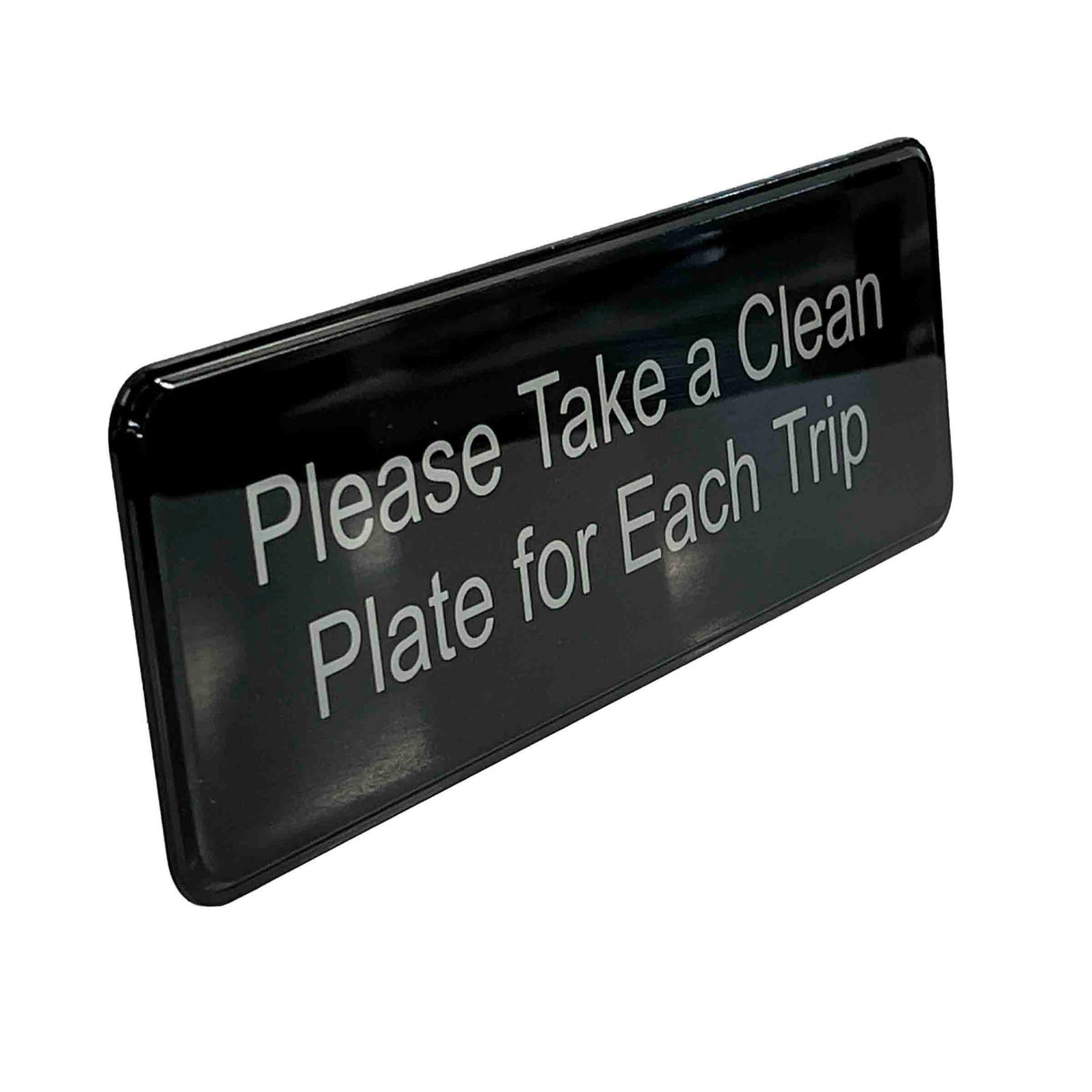 A rectangular black TableCraft Products clean plate sign with white text reads, Please Take a Clean Plate for Each Trip. The sign has rounded edges, a glossy finish reflecting light, and features self adhesive backing for easy mounting.