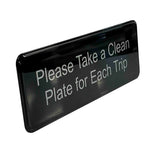 A rectangular black TableCraft Products clean plate sign with white text reads, Please Take a Clean Plate for Each Trip. The sign has rounded edges, a glossy finish reflecting light, and features self adhesive backing for easy mounting.