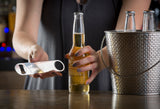 A person in a dark shirt holds a bottle of beer and a stainless steel pocket bottle opener. Next to them, a silver ice bucket chills three more bottled beers, evoking the relaxed vibe of a bar or casual gathering.