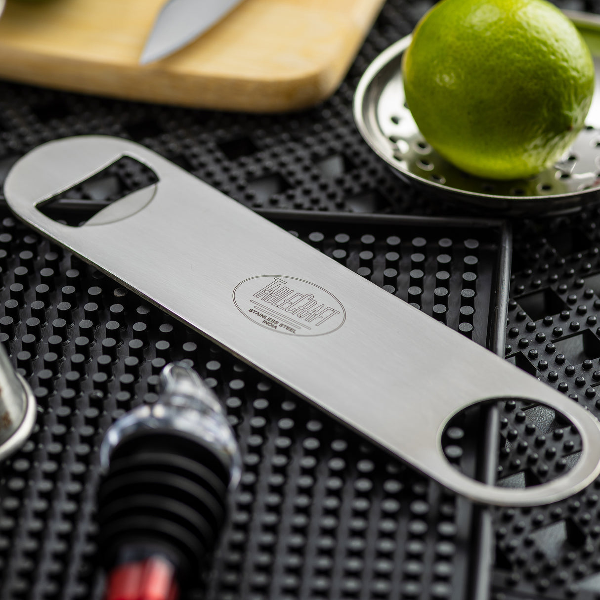 A stainless steel pocket bottle opener lies on a black textured bar mat, surrounded by a lime, a metal citrus zester, a bottle stopper, and a knife on a wooden board in the background. The dishwasher safe opener has two round holes and engraved text.
