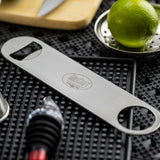 A stainless steel pocket bottle opener lies on a black textured bar mat, surrounded by a lime, a metal citrus zester, a bottle stopper, and a knife on a wooden board in the background. The dishwasher safe opener has two round holes and engraved text.