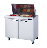 The Blue Air Mega Top sandwich prep table features a stainless steel build, two doors, a glass lid displaying assorted toppings in trays, four caster wheels, and a digital temperature display on the right side.