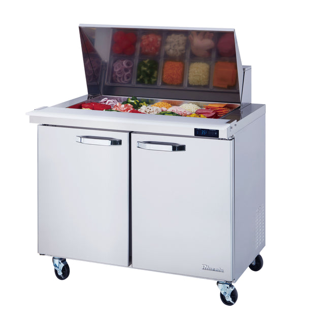 The Blue Air Mega Top sandwich prep table features a stainless steel build, two doors, a glass lid displaying assorted toppings in trays, four caster wheels, and a digital temperature display on the right side.
