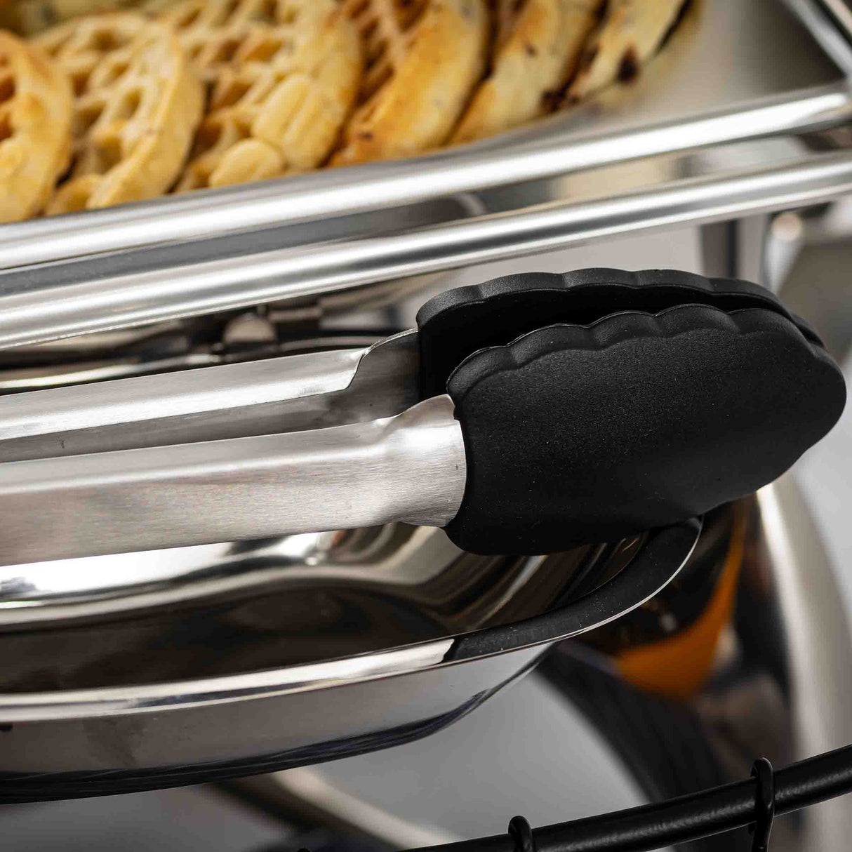 A close-up of stainless steel tongs with black silicone grips resting on the edge of a tray filled with round, waffle-shaped pieces of food. The clean, shiny setup suggests a buffet or catering setting using quality, heat resistant tongs.