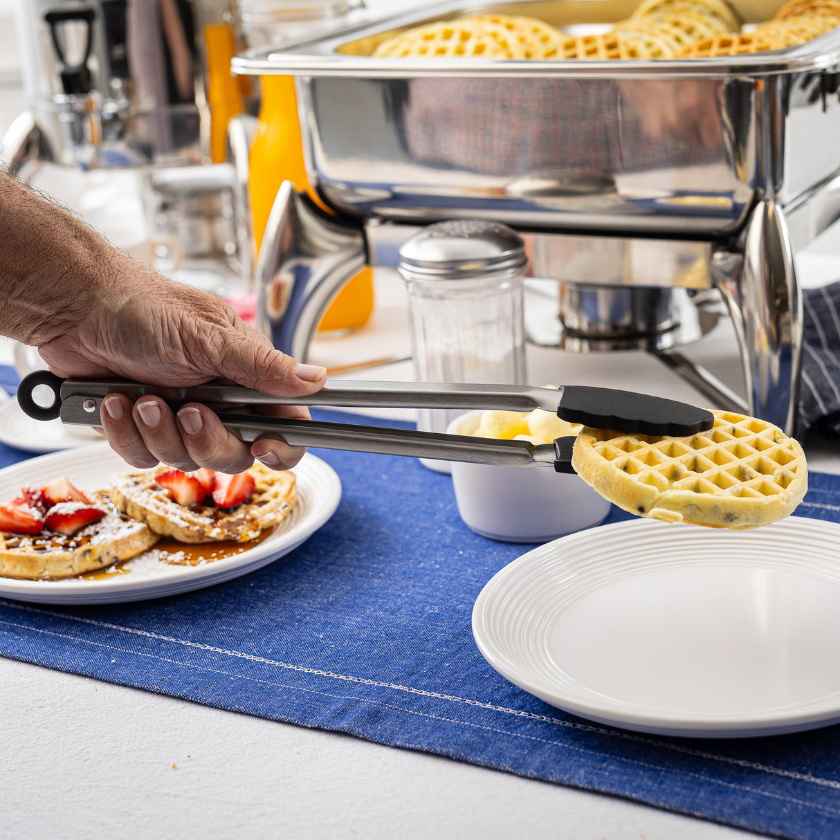 A hand uses stainless steel tongs to place a small round waffle onto a white plate. Nearby, plates with waffles topped with powdered sugar and strawberries sit on a blue table runner, alongside a chafing dish of waffles and a glass jar of sugar.