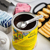 An opened can of Café Bustelo Brazil ground coffee with a stainless steel coffee scoop on top. Nearby are blue and pink sweetener packets, a coffee cup, and a tray of ladyfingers set on a white tablecloth.