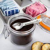 An open glass jar filled with ground coffee sits on a white surface, with a stainless steel teaspoon inside. Nearby, metal containers hold blue and pink sweetener packets, and part of a pastry is visible in the foreground.