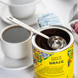 An opened can of Café Bustelo Brazil coffee with a stainless steel coffee scoop inside is shown. Next to it is a cup of black coffee in a white cup and saucer, and a container holding blue and pink sweetener packets on a white surface.