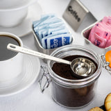 A glass jar of ground coffee with a stainless steel 2 tablespoon Coffee Scoop inside sits on a table. Nearby are a cup of black coffee, blue and pink sweetener packets in metal containers, and part of a plate with pastries on a white tablecloth.