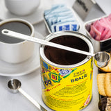 A can of Café Bustelo Brazil ground coffee is open, with a long handled stainless steel measuring spoon resting on top. Nearby are a cup of black coffee, sweetener packets in metal trays, another scoop, cookies, and chocolate on a white surface.