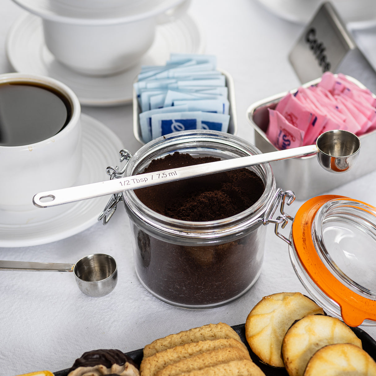 A jar of ground coffee with a long handled stainless steel measuring spoon on top sits on a table. Surrounding it are coffee cups, blue and pink sweetener packets, assorted cookies, and a partially open jar lid with an orange rim—perfect for a coffee break.