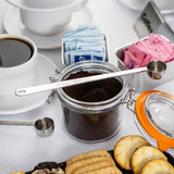 A jar of ground coffee with a long handled stainless steel measuring spoon on top sits on a table. Surrounding it are coffee cups, blue and pink sweetener packets, assorted cookies, and a partially open jar lid with an orange rim—perfect for a coffee break.