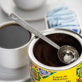 A long handled stainless steel measuring spoon rests inside an open can of Café Bustelo ground coffee. Next to it is a white cup filled with black coffee on a saucer, and a container of blue sugar packets, all set on a bright, white surface.