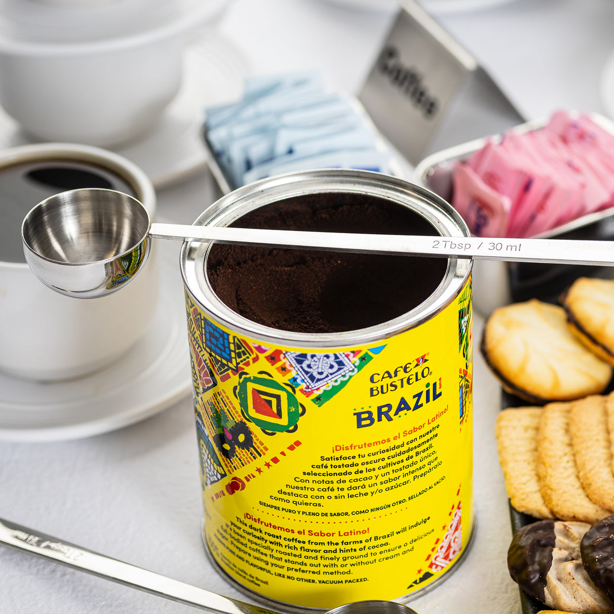 A can of Café Bustelo Brazil ground coffee with a colorful label is open, showing coffee grounds inside. A long handled stainless steel measuring spoon rests on top. Nearby are coffee cups, sugar packets, and cookies on a white surface.