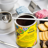 A can of Café Bustelo Brazil ground coffee with a colorful label is open, showing coffee grounds inside. A long handled stainless steel measuring spoon rests on top. Nearby are coffee cups, sugar packets, and cookies on a white surface.