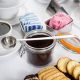 A jar of ground coffee with a long handled stainless steel measuring spoon rests on top. Nearby are blue and pink sweetener packets in trays, a cup of black coffee, stacked white cups, and assorted cookies, all on a white tablecloth.