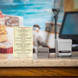 A restaurant counter with a daily specials menu in an acrylic menu holder, a Reserved sign, and a receipt printer. The blurred background features a mural with a figure by the sea. The beige countertop highlights the signs and printer up front.