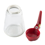 A clear teardrop glass jar is upside down next to a red plastic jar opener with a handle and a rotating metal band. The opener, designed for ease, helps twist lids open. Both items are placed on a white background.