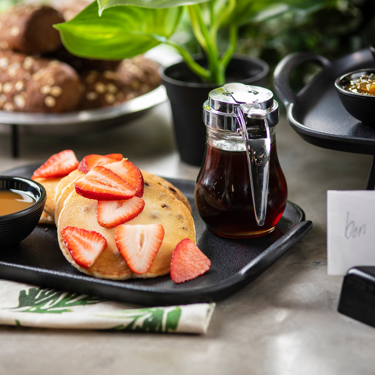 Two pancakes topped with fresh strawberry slices are served on a black plate, accompanied by a teardrop glass syrup dispenser and a small bowl of sauce. Bread rolls and a napkin with a leaf design complete the stylish scene.