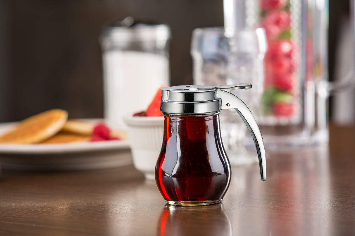 A teardrop glass syrup dispenser with a metal lid and handle sits on a brown table. In the blurred background are pancakes, a cup, a sugar shaker, and a glass pitcher with red fruit and water—perfect for any breakfast table.