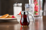 A teardrop glass syrup dispenser with a metal lid and handle sits on a brown table. In the blurred background are pancakes, a cup, a sugar shaker, and a glass pitcher with red fruit and water—perfect for any breakfast table.