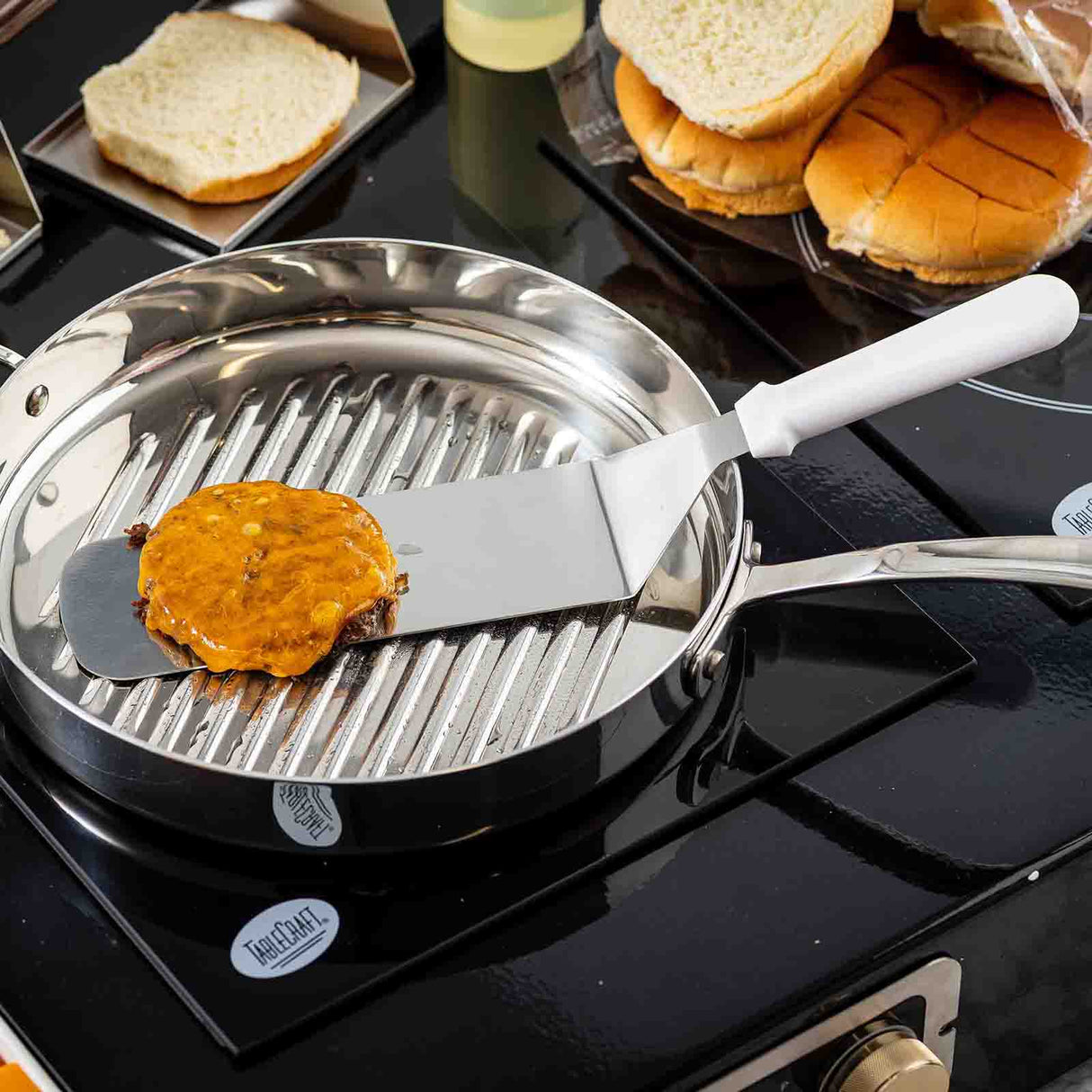 A stainless steel Food Turner with a dishwasher safe blade lifts a cooked hamburger patty with melted cheese from a ridged pan on the stovetop. Sliced and whole buns sit in the background, ready for delicious sandwich assembly.