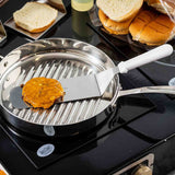 A stainless steel Food Turner with a dishwasher safe blade lifts a cooked hamburger patty with melted cheese from a ridged pan on the stovetop. Sliced and whole buns sit in the background, ready for delicious sandwich assembly.