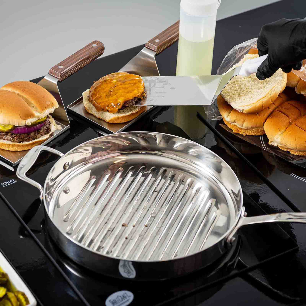 A stainless steel grill pan sits on a stovetop. Next to it, a hand in a black glove uses a dishwasher safe Food Turner with a stainless steel blade to place a cheeseburger patty onto an open bun. A squeeze bottle and pack of buns are nearby.