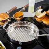 A stainless steel grill pan sits on a stovetop. Next to it, a hand in a black glove uses a dishwasher safe Food Turner with a stainless steel blade to place a cheeseburger patty onto an open bun. A squeeze bottle and pack of buns are nearby.