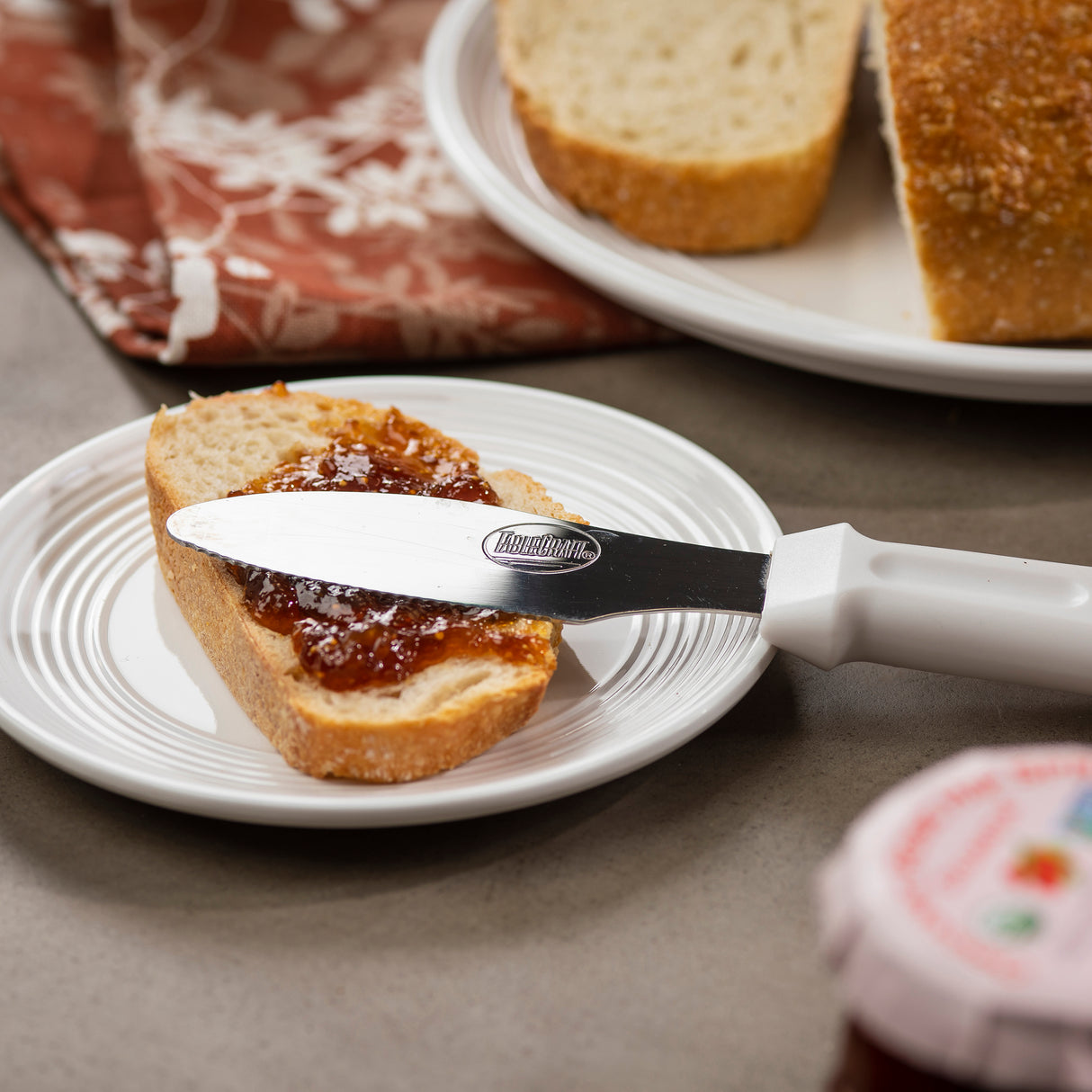 A slice of bread with jam spread on it sits on a white plate. A stainless steel sandwich spreader rests on the bread. In the background, there is more bread on a plate, a patterned cloth, and a partially visible jar with a red label.