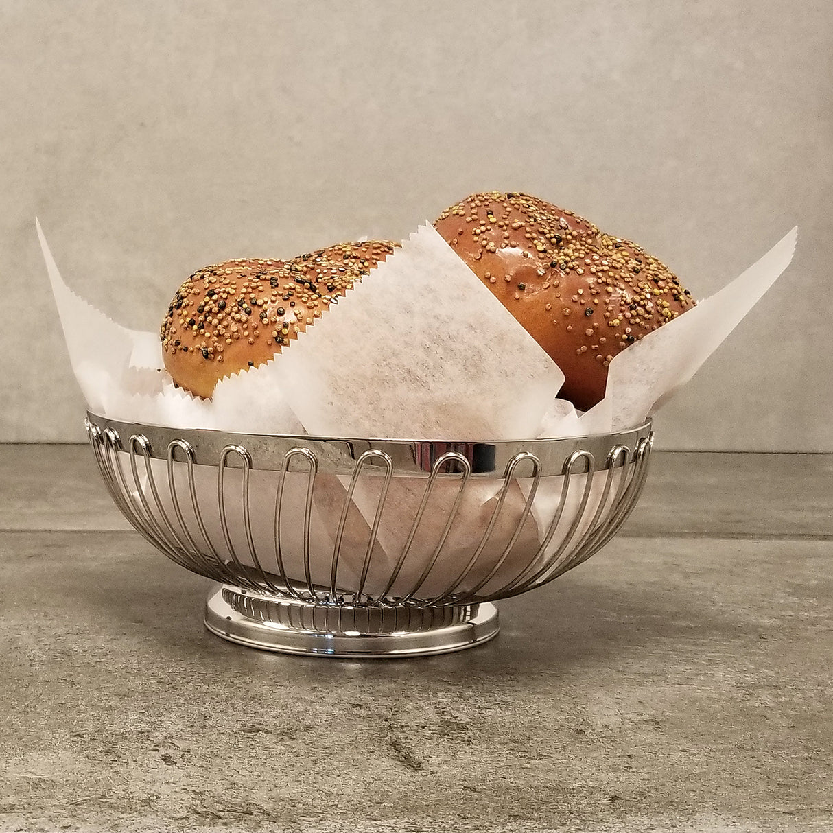 A chrome plated Chalet Basket from TableCraft Products, lined with parchment paper, holds two seeded burger buns with colorful seeds, resting on a gray stone surface against a plain gray background.