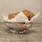 A chrome plated Chalet Basket from TableCraft Products, lined with parchment paper, holds two seeded burger buns with colorful seeds, resting on a gray stone surface against a plain gray background.