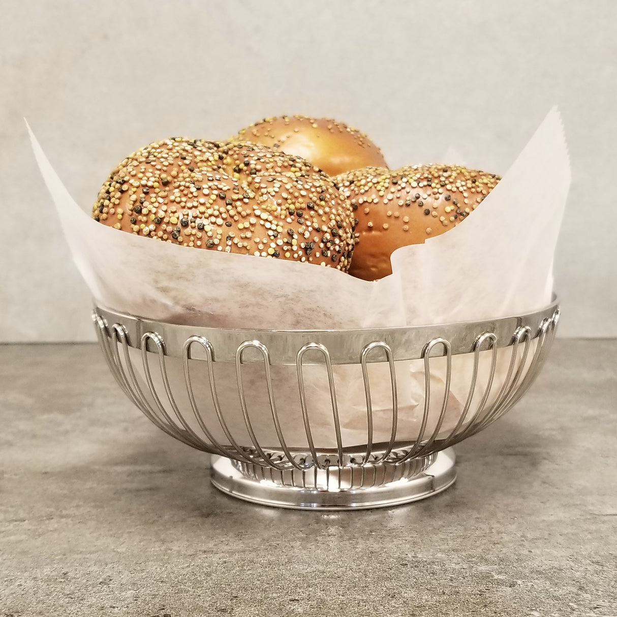 A chrome plated Chalet Basket from TableCraft Products, lined with white parchment paper, holds three round, golden brown buns topped with multicolored seeds. The basket sits on a gray surface against a light gray background.