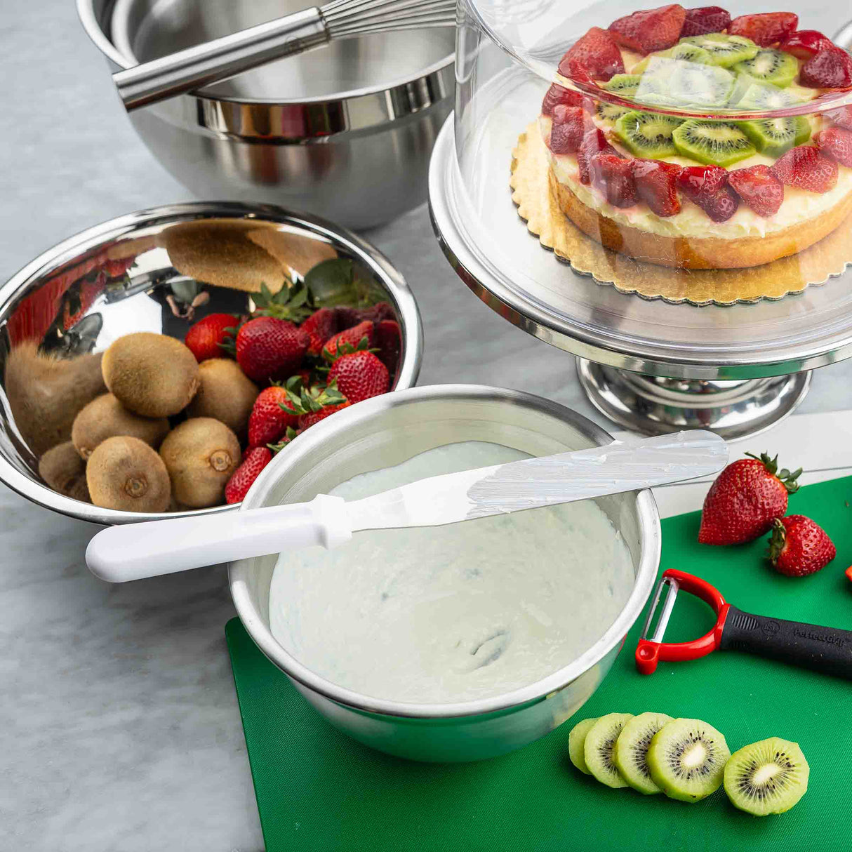 A kitchen scene with metal bowls, whole kiwis, strawberries, a cake topped with sliced kiwi and strawberries under a glass dome, a bowl of cream with an icing spatula, a green cutting board with sliced kiwi, a strawberry, and a red peeler.