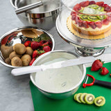 A kitchen scene with metal bowls, whole kiwis, strawberries, a cake topped with sliced kiwi and strawberries under a glass dome, a bowl of cream with an icing spatula, a green cutting board with sliced kiwi, a strawberry, and a red peeler.