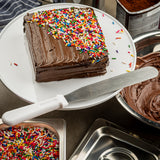 A square chocolate cake with chocolate frosting and rainbow sprinkles sits on a white cake stand. An icing spatula with a stainless steel blade and white handle rests nearby, and bowls of frosting and rainbow sprinkles surround the cake.