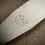 Close-up of a metallic knife blade with an oval-shaped “KÜCHEN” logo near the handle. The brushed stainless steel blade appears clean and well-crafted, lying on a textured surface with parallel lines engraved beneath the text.