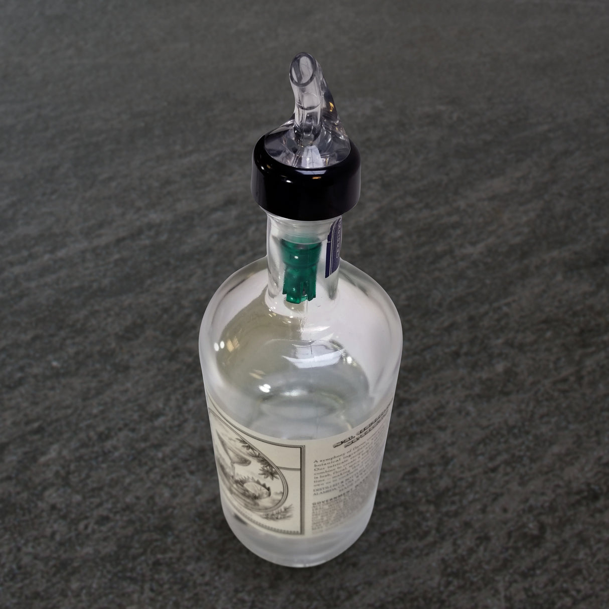 A clear glass bottle with a white label and black-and-white design sits on a gray textured surface. The bottle features a TableCraft 3/4 oz pourer and black Proper Pour spout, with a green internal stopper visible at the neck.