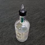 A clear glass bottle with a white label and black-and-white design sits on a gray textured surface. The bottle features a TableCraft 3/4 oz pourer and black Proper Pour spout, with a green internal stopper visible at the neck.