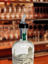 A clear liquor bottle with a TableCraft 3/4 oz. pour spout is shown in focus. The bottle is partially filled and sits on a bar counter, while shelves stocked with various bottles are blurred in the background.