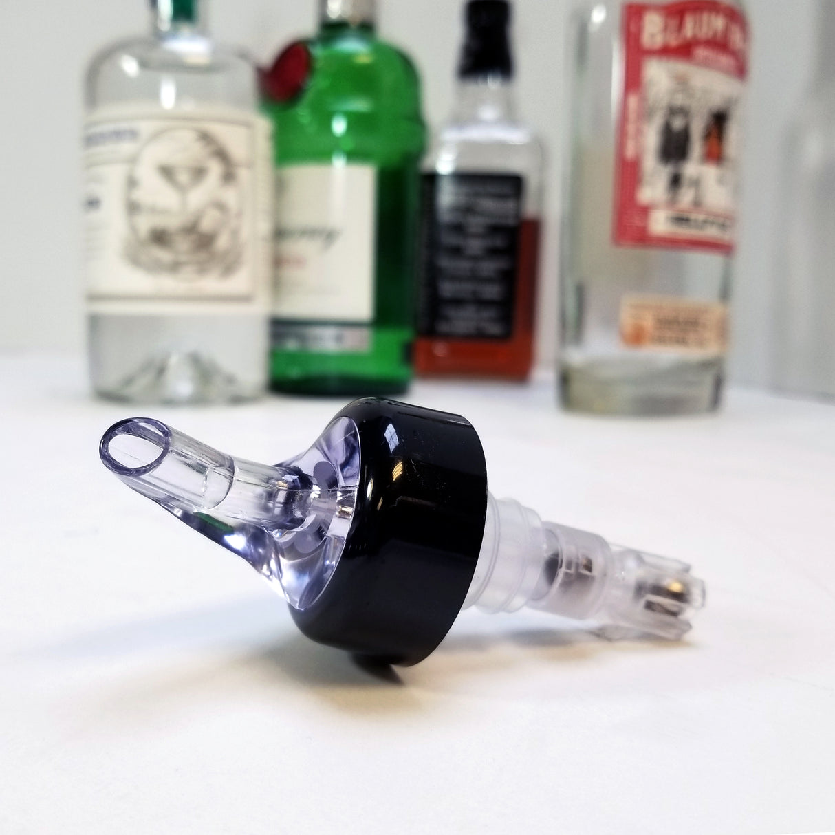 A clear plastic 1-1/4 oz pourer with a black collar lies on a white surface. In the blurred background, liquor bottles with colored labels and caps suggest a bar or home bartending setup—ideal for achieving the Proper Pour every time.