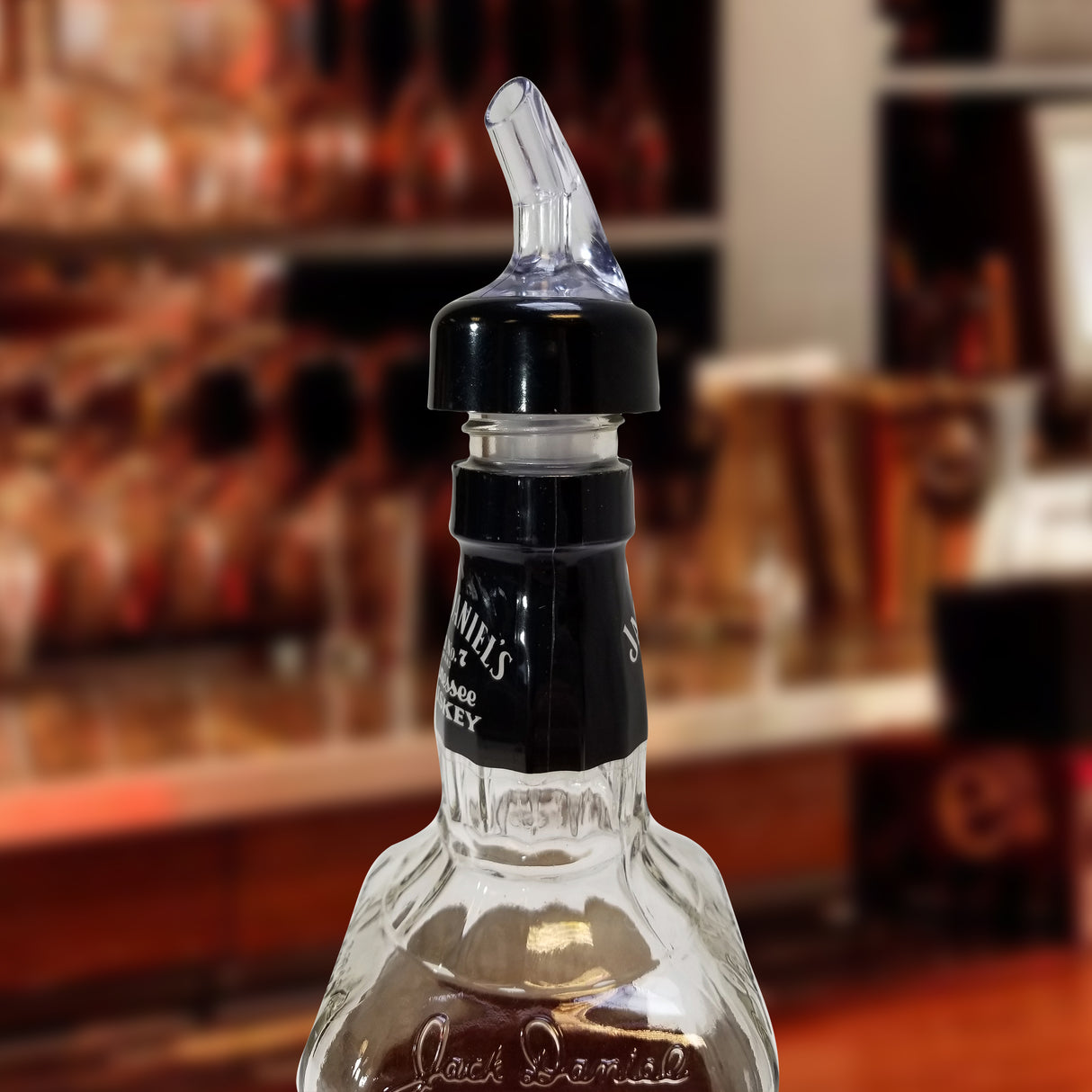 A close-up of a glass whiskey bottle with a black screw cap and a TableCraft Products Proper Pour 1-1/4 oz pourer. The blurred background reveals shelves with bottles, suggesting a bar or liquor store setting.
