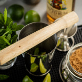 A natural wood TableCraft muddler rests on a metal shaker containing lime wedges. Fresh mint, whole limes, a jar of brown sugar, a jigger, and rum are arranged nearby, suggesting the makings of a classic mojito cocktail.