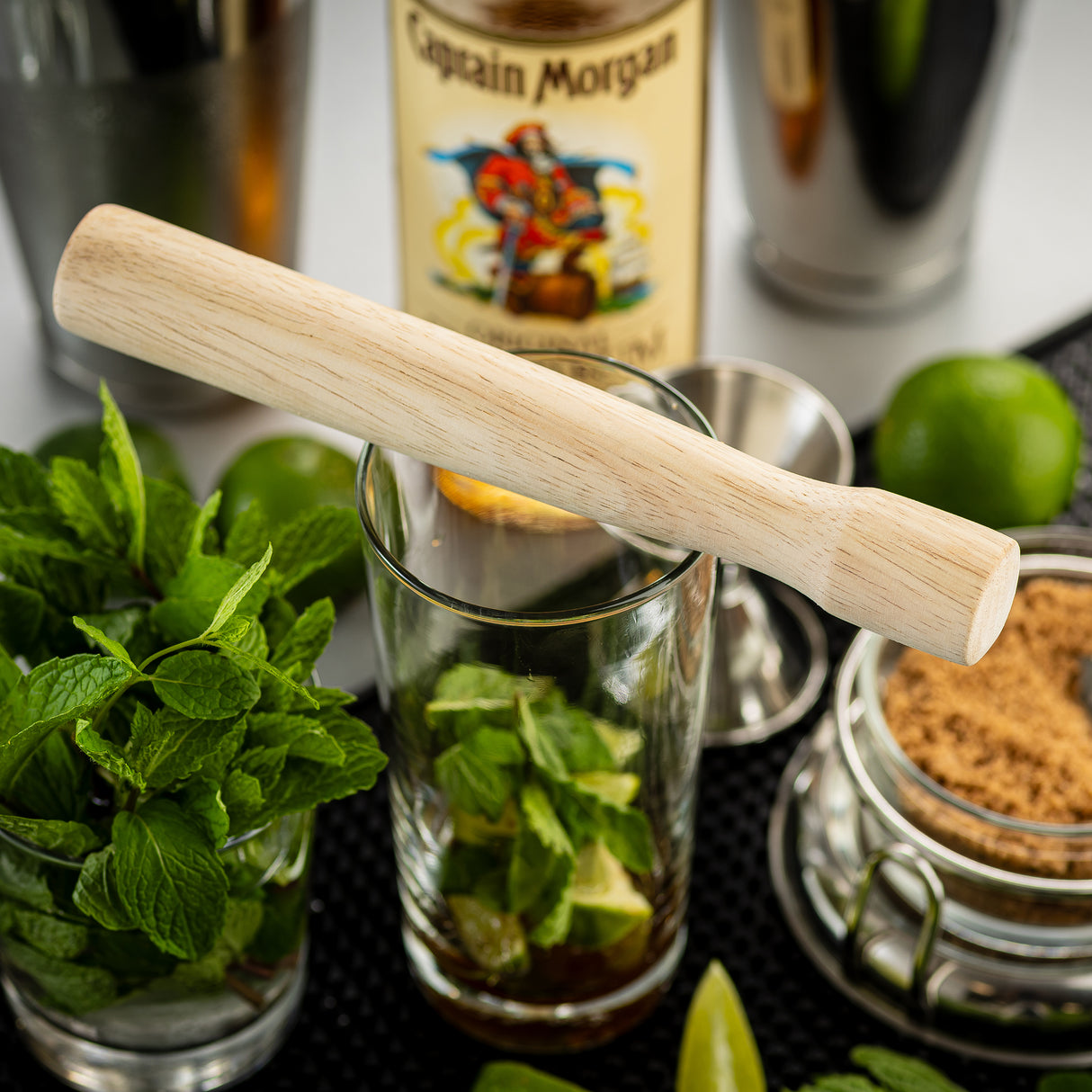 A TableCraft natural wood muddler rests across a glass filled with mint leaves. Nearby are a jar of brown sugar, whole and sliced limes, fresh mint, Captain Morgan rum, a cocktail shaker, and bar tools arranged on a bar mat.